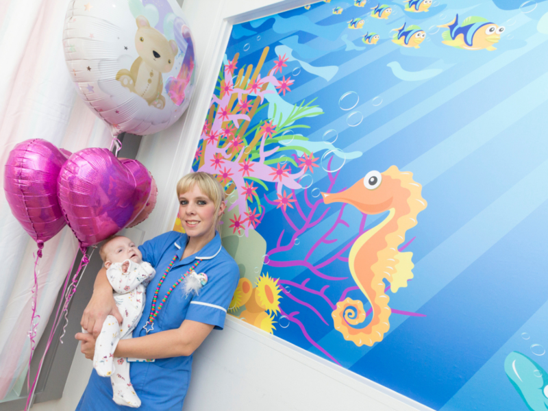 A nurse holds a baby in front of the a seahorse mural